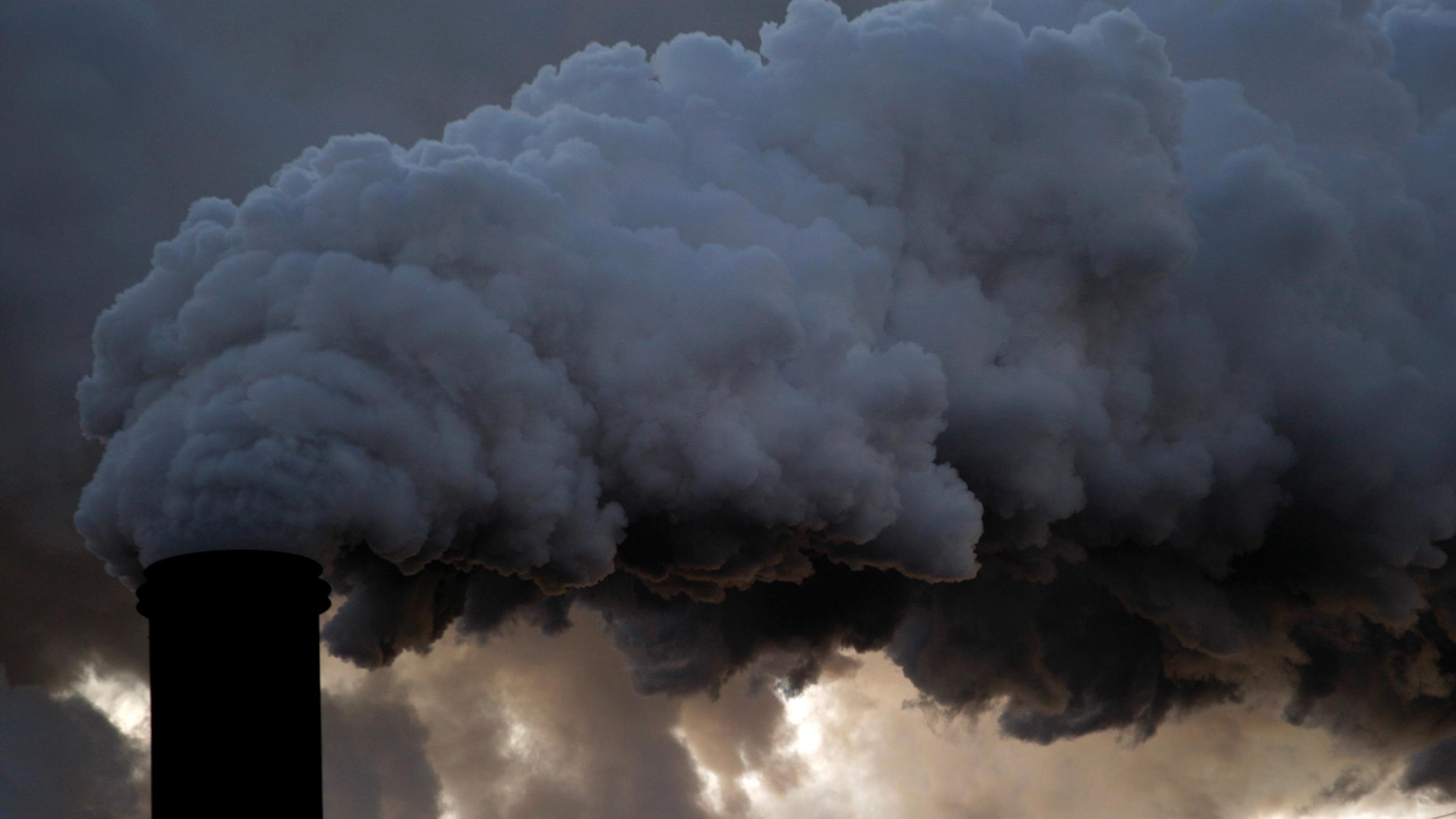 The image shows a large smokestack belching out black billows and blocking the cloudy sky.