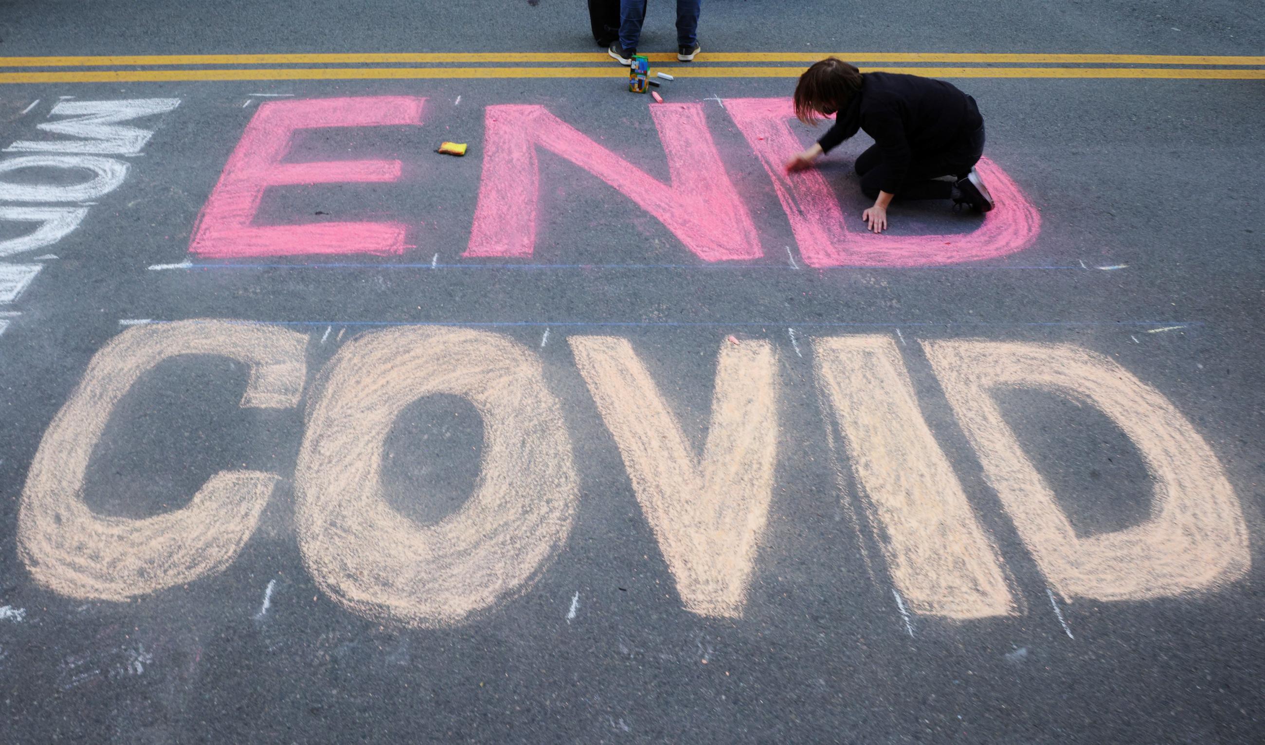 Demonstrators write "End COVID" in pink and yellow sidewalk chalk on the street outside Moderna headquarters, pushing for the company to share its COVID vaccine technology, in Cambridge, Massachusetts, on April 28, 2022.